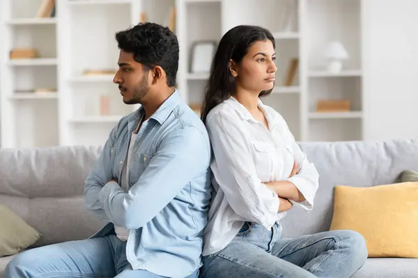 Young indian couple sits back-to-back on sofa, surrounded by a tense atmosphere, visibly upset and offended following a heated quarrel in their homey living space