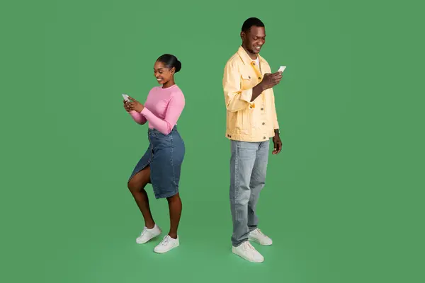 Gadgets addiction concept. Positive smiling stylish young black man and woman using cell phones isolated on green studio background, chatting, scrolling. Dating mobile application