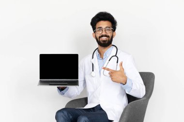 Genç Hintli bir doktor, dizüstü bilgisayarını boş bir ekranla sunuyor, beyaz bir arkaplana karşı sandalyede oturuyor, dijital sağlık iletişimini, teletıp, kopyalama alanını, taklit alanını sembolize ediyor,