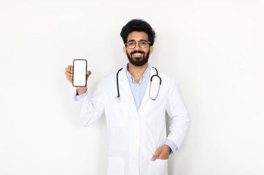 Genç Hintli bir doktor, kendine güvenen bir akıllı telefonu boş bir ekranla sunuyor, beyaz bir arka plana karşı duruyor, dijital sağlık iletişimini, teletıp, kopyalama alanını, taklit alanını sembolize ediyor,