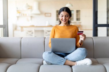 Gülümseyen Hintli genç bir kadın evdeki koltukta otururken internetten bilgisayar ve kredi kartıyla alışveriş yaparken mutlu bir milenyum kadını internetten satın almanın keyfini çıkarıyor.
