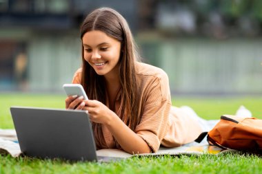Gülümseyen genç bir kız öğrenci bilgisayarında ve telefonunda aynı anda boş bir üniversitede geziniyor, çimlere uzanıyor, çevrimiçi bir derse katılıyor ve çalışma notlarıyla ilgili grup sohbetine katılıyor.