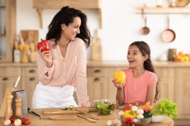 Neşeli anne ve küçük kız taze organik sebzelerden salata yaparken eğleniyorlar, yemek pişiriyorlar ve gülüyorlar, modern mutfaktaki masanın yanında dolmalık biber tutuyorlar.