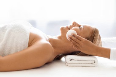 Profesyonel kozmetik uzmanı cildini dikkatlice orta yaşlı, sakin bir kadından temizliyor. Lüks güzellik salonunda cilt bakımı sırasında pamuk pedler kullanıyor. Gözleri kapalı gülümseyen bir kadın.