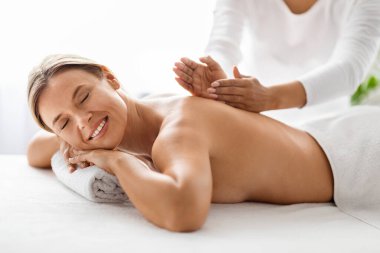 Masör elleri masaj masasında dinlenen orta yaşlı güzel bir kadın lüks spa salonunda terapi seansı düzenlerken sakin, gülümseyen bir kadın omurga tedavisi görüyor.