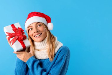 Noel Baba şapkası takan heyecanlı beyaz kadın hediye kutusunu tutuyor ve mavi arka planda boş alana bakıyor, tahmin et içinde ne var. Tatil havası.