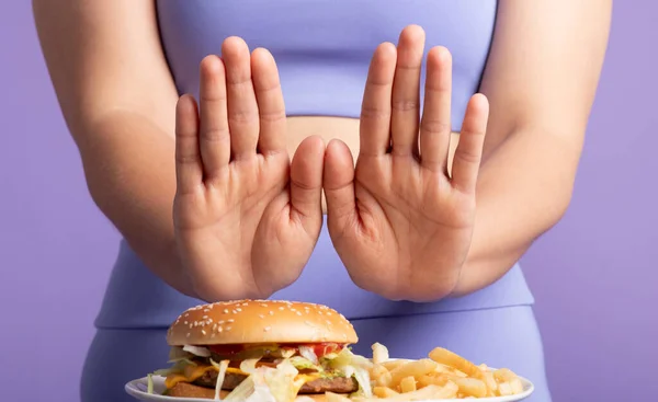 Sporty woman confidently showing stop gesture toward tempting burger and fries, promoting healthy dietary choice, standing over purple studio background, closeup. Weight loss, body care, diet