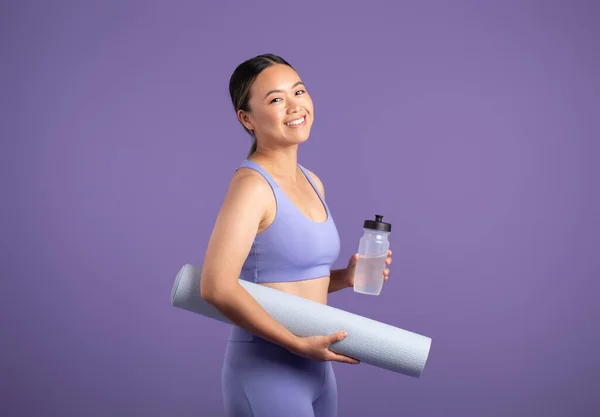 Energetic Asian woman in sportswear, holding yoga mat and bottle of water, preparing for refreshing workout, posing against purple studio background