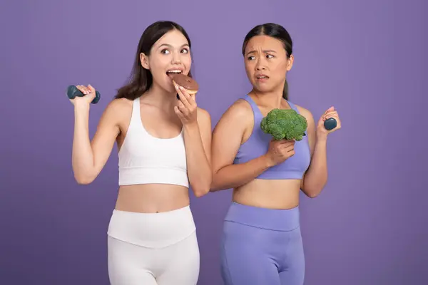 Asian lady holding broccoli, covets a donut being eaten by caucasian woman, ladies in sportswear, posing on purple studio background, illustrating a dietary dilemma
