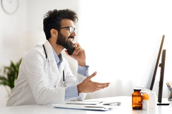 Handsome Indian man doctor, immersed in work, juggling tasks between computer and phone, giving remote consultation, sits at a clinic desk, representing a bustling healthcare environment, copy space
