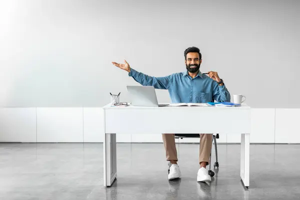 Handsome indian businessman sitting at desk with laptop and pointing aside at free place with hand. Male entrepreneur in formalwear showing space for your advertisement or text