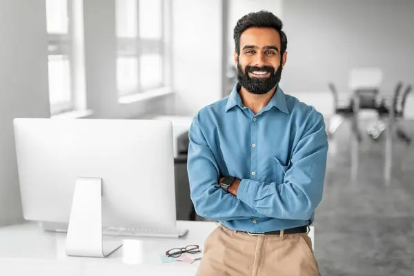 Happy indian businessman standing by his workplace with folded arms, smiling at camera, planning investment, thinking of new startup or project, enjoying business success