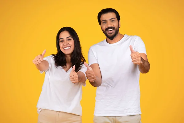 Enthusiastic young arab couple show thumbs up, isolated on yellow studio background. Expressions and gestures show approval and positivity, making atmosphere fun and uplifting