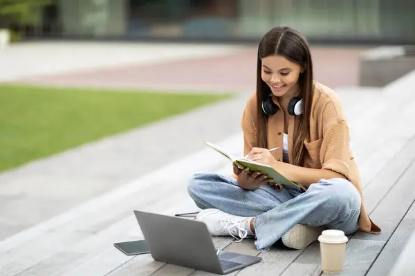 In a silent university space, a pleased young woman student operates her laptop, possibly engaged in an educational podcast or webinar, eyes locked on the screen, taking notes in notepad, copy space