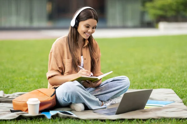 A young woman student at an empty university campus is smiling, taking notes at notepad, using laptop and headphones, enjoying online lecture, attending webinar, online class, copy space