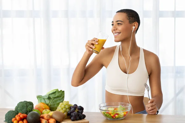 Smiling slim young latin lady in sportswear drink orange juice, eat salad, listen music in kitchen interior with fresh vegetables. Diet, weight loss, healthy food, proper nutrition at home