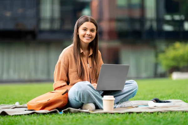笑顔の女性学生が大学のキャンパスでノートパソコンを使っている 緑の芝生に座って 彼女はタイプし 時には彼女のスクリーンに焦点を当てながら活発なキャンパスの雰囲気を吸収し 周りを見回します — ストック写真