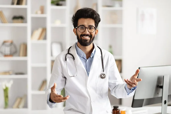 Portrait of cheerful eastern man doctor gesturing and smiling while working at clinic in his cozy office, physician wearing white medical coat standing next to desk with computer, have appointment