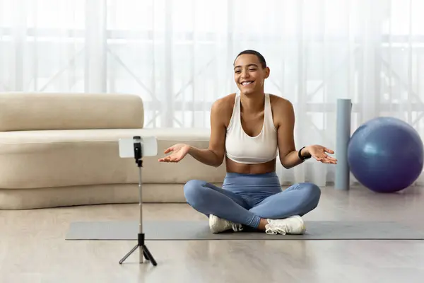 Positive confused millennial latin lady blogger in sportswear sit on floor, shooting video fitness blog on phone in living room interior. Weight loss and sports at home, fitness