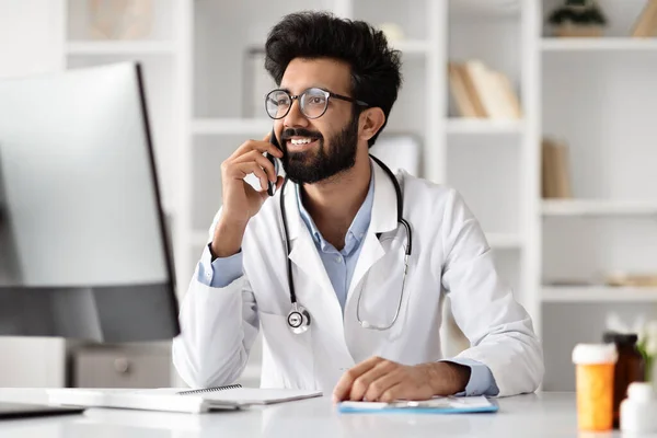 Busy young Indian healthcare professional, engaging in telecommunication, balances computer work and a phone call with remote patient at a clinic, doctor portraying efficient work management