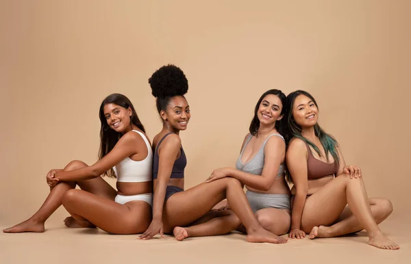 Embracing diversity and beauty, four multiracial women gracefully sit on the studio floor, each manifesting unique allure, smiling at camera, full length