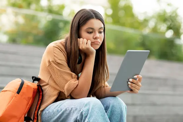 屋外でデジタルタブレットを使った若い女性学生 大学のキャンパスや公共公園だけに座って パッドスクリーンを見たり 退屈したり孤独を感じたり 宿題に困難を感じたりする — ストック写真