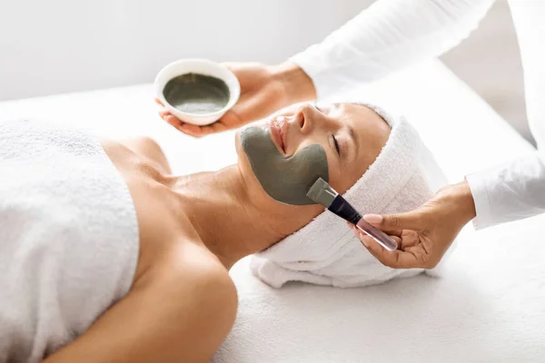 In luxury spa salon beautician applying nourishing clay mask to middle aged woman face, offering soothing, revitalizing skincare ritual to attractive calm mature lady lying on table, closeup shot