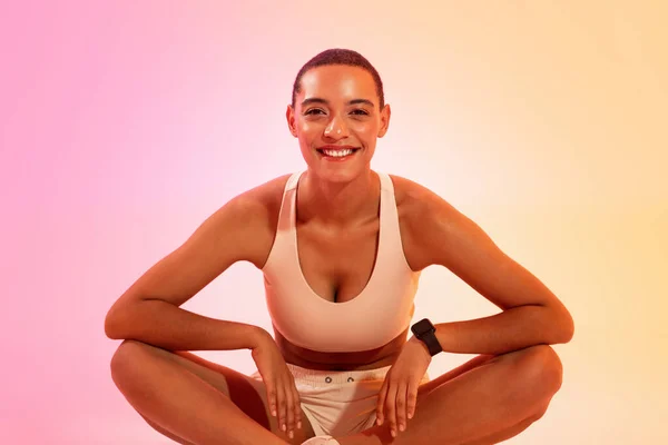 Smiling tired pretty young sweat latin woman in sportswear sitting, resting from workout on neon pink studio background. Fitness, sport, health care and weight loss