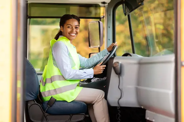 Professions Concept. Portrait Of Young Black Female Driver Posing In Bus, Happy African American Woman Wearing Uniform Holding Steering Wheel And Smiling At Camera, Enjoying Her Job, Free Space