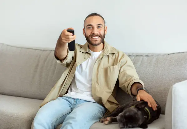Watching TV at home. Happy millennial man having rest, switching channels with remote controller and smiling, empty space, enjoying his weekend, watching TV in living room, petting dog