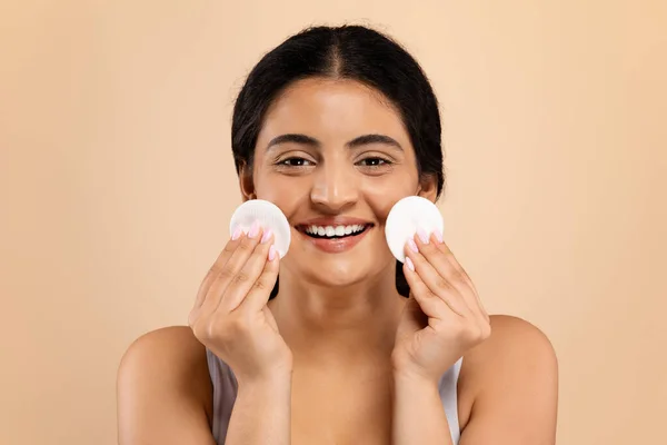 Skincare Concept. Happy Beautiful Indian Female Cleaning Face With Two Cotton Pads, Attractive Eastern Woman Enjoying Beauty Routine, Standing Isolated Over Beige Background, Copy Space