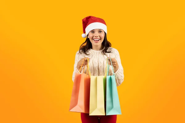 Xmas and winter holidays shopping. Positive young woman in Santa Claus hat holding Christmas gift bags over yellow studio background. New Year celebration and sales concept