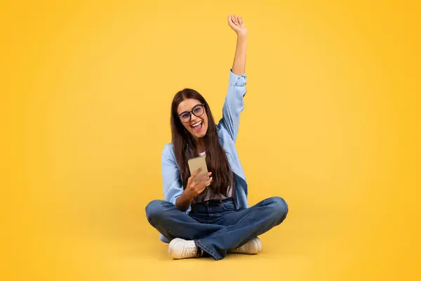 满意的戴眼镜的女学生看着电话 庆祝胜利 坐在地板上 被黄色背景隔离 设备方面的胜利 销售和好消息 — 图库照片