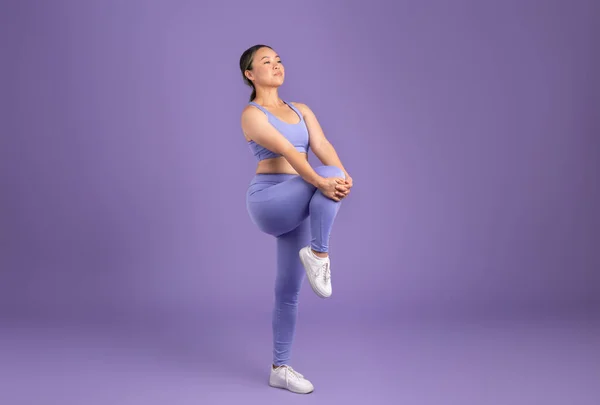Young asian woman in sportswear warming up and stretching legs, on purple studio background, showing active moment, full length shot, free space. Sports, weight loss, fitness and body care