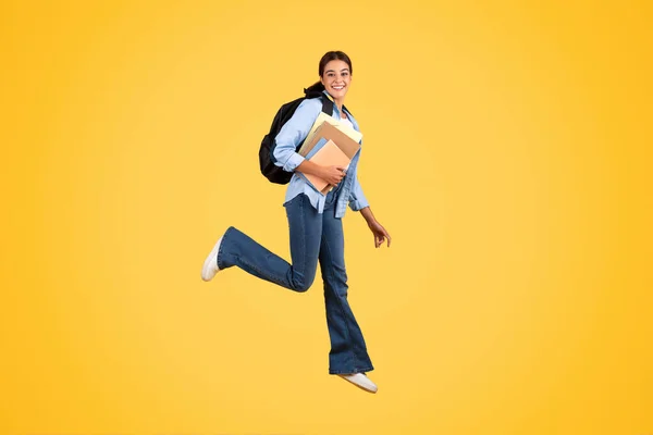 快乐的年轻欧裔女士 背着背包 耳机和书本跳跃 空气冻结 享受宁静的学生生活 在黄色的工作室背景下与世隔绝 放松和有趣 — 图库照片