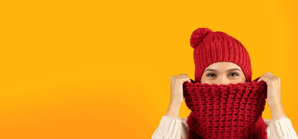 赤い編まれた帽子を身に着けている若い女性は黄色いスタジオの背景の上の冬の冷たいから隠れ カメラ パノラマ 自由なスペースを見ている暖かいスカーフの表面をカバーします — ストック写真
