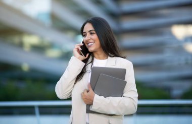 İş İletişim Konsepti. Cep telefonuyla konuşan, gülümseyen, şehir dışında duran, elinde dijital tablet ve dizüstü bilgisayarla gezen mutlu iş kadını.