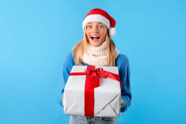 Harika bir Noel hediyesi. Noel Baba şapkalı heyecanlı bayan elinde Noel hediyesi kutusu ve mavi stüdyo arka planında ağzı açık kameraya bakıyor. Kış bayramı kutlaması ve hediye konsepti