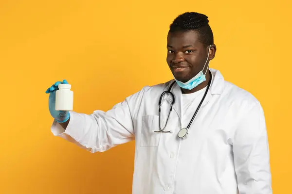 Smiling african american therapist guy shows medical jar of pills in hand on yellow studio background. Doctor giving medicines treatment advice, pharmaceutics choice and offer concept