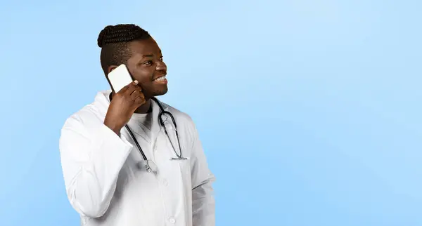 Call Your Doctor. Cheerful black general practitioner man talking on cellphone looking aside, standing over blue studio background. Healthcare and communication. Panorama with copy space