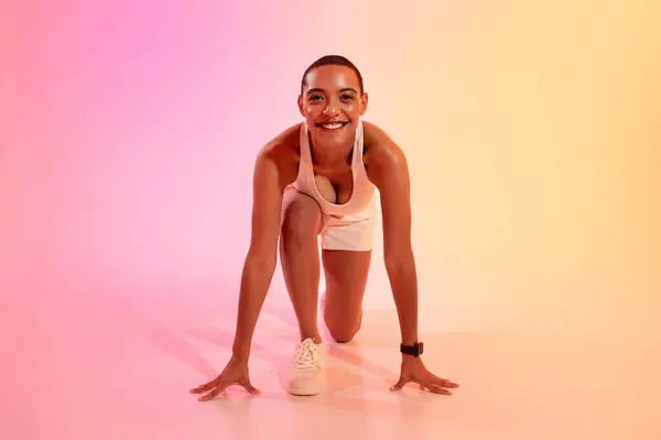 Happy young latin woman in sportswear stands at start, ready to run on neon pink studio background. Competition, jogging, professional sports, health care, weight loss
