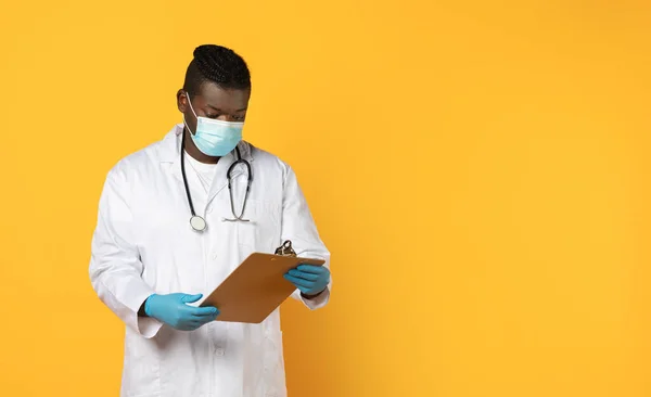 Black man doctor in face mask and white coat with tablet clipboard on yellow studio background. Professional medical diagnostics, health care treatment and medicine concept. Copy space