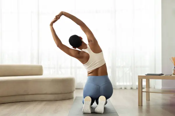 Young slim latin woman in sportswear doing body stretching on mat in living room interior, back. Health care, weight loss and sports at home, fit lifestyle and vitality