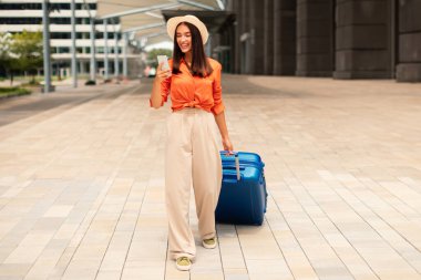 Turizm ve Gadget 'lar. Akıllı telefon kullanan turist kadın çantasıyla ayakta duruyor ve dışarıdaki Modern Havaalanı Terminali 'nde seyahat biletleri ayırtıyor. Tam Uzunluk Çekimi