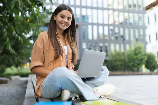 一个快乐的女学生一个人坐在大学的长椅上 专心致志地在笔记本电脑上打字 也许是在空旷的环境中完成作业 对着相机微笑 抄袭空间 — 图库照片