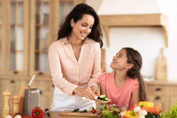 Young mommy and daughter preparing fresh salad for dinner in the kitchen. Family activities and healthy culinary habits. Mom adding cut vegetables to bowl smiling to kid girl