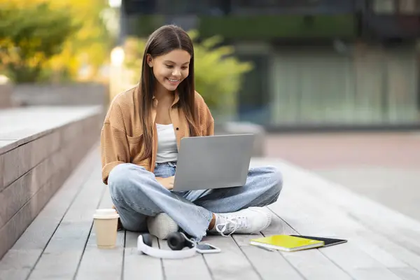 ポジティブな若い女性学生は 空のキャンプスベンチにソロに座って ノートパソコンとやり取りしたり プロジェクトの宿題を提出したり メールを送信したり レッスン後に休憩しながらオンラインで作業したりします — ストック写真