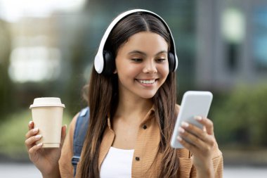 Neşeli genç bayan öğrenci dışarıda müzik dinliyor, kablosuz kulaklık ve akıllı telefon kullanıyor, paket kahve içiyor, üniversitede tatildeyken favori podcastın tadını çıkarıyor.