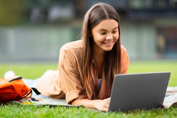 広い笑顔で 若い女性の学生は 空の大学のスペースで芝生に一人で位置し 彼女のラップトップ上のプロジェクトやオンラインの割り当てを通じて熱心に進み スペースをコピーします — ストック写真
