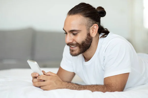 Cheerful handsome caucasian man in pajamas lying on bed at home, using smartphone and smiling, checking social media while chilling alone at weekend, copy space. Leisure and gadgets